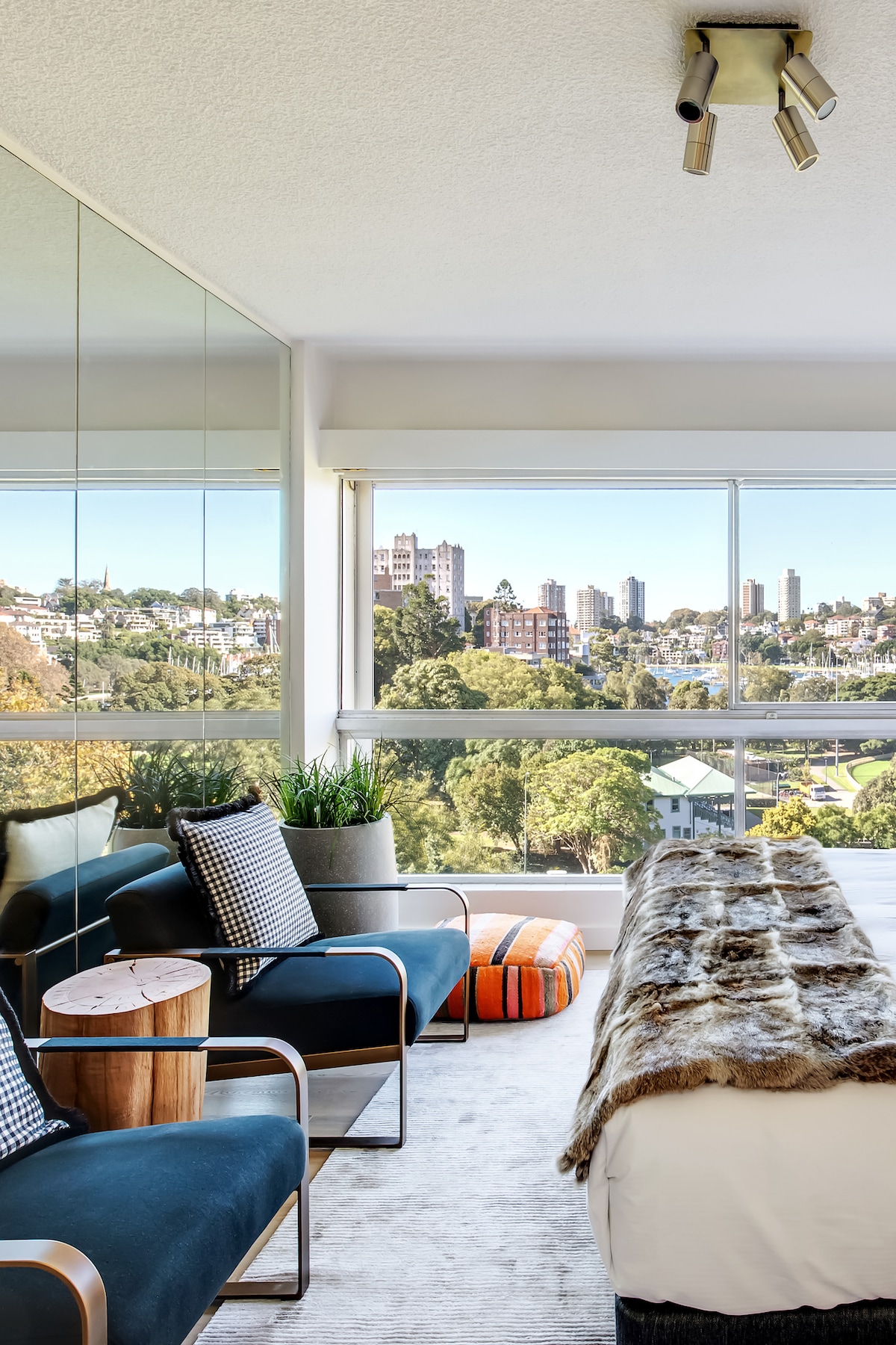 A bright bedroom offers a spacious queen bed adorned with a soft throw. Two comfortable chairs, accented with checkered cushions, flank a wooden side table. Floor-to-ceiling windows provide a clear view of the city skyline, with lush treetops visible in the foreground.