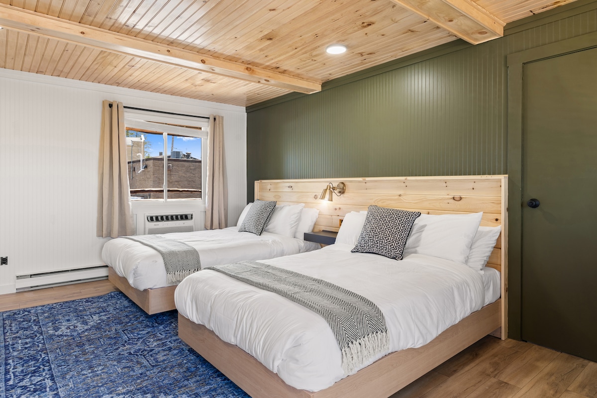 A well-lit bedroom features two spacious beds with soft white linens and decorative throws. Natural wood accents and a vibrant blue rug add warmth. Large windows allow sunlight to fill the space, while neutral-colored curtains provide privacy.