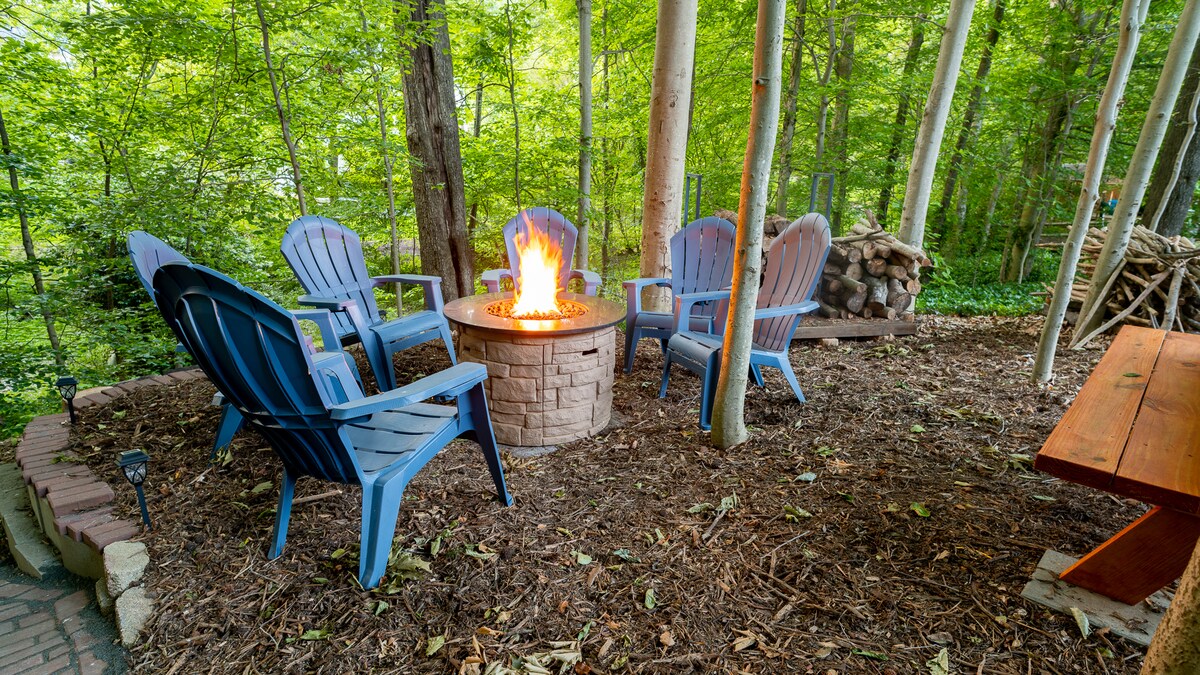 A cozy outdoor fire pit is surrounded by several light blue Adirondack chairs, set in a natural clearing among trees. The warm glow of the flames lights the area, with a stack of firewood nearby and a picnic table in the background.