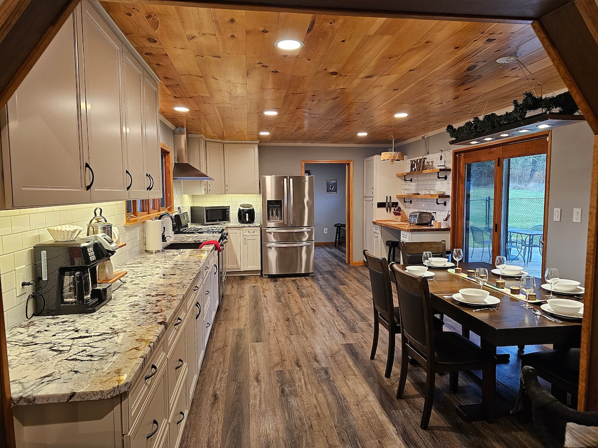 A renovated kitchen is highlighted by modern appliances and natural wood accents. A spacious dining table is set for a meal against a backdrop of large windows that provide views of the outdoor space. Ample counter space and cabinetry enhance functionality.
