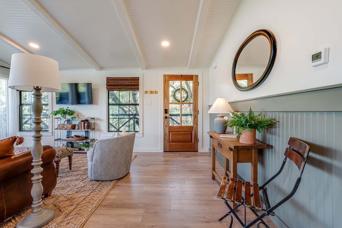 A well-lit living area showcases a comfortable seating arrangement with a leather sofa and a modern accent chair. A wooden table and decorative lamp are positioned next to a window, adding functionality. A large mirror and a television provide balance and enhance the inviting environment.