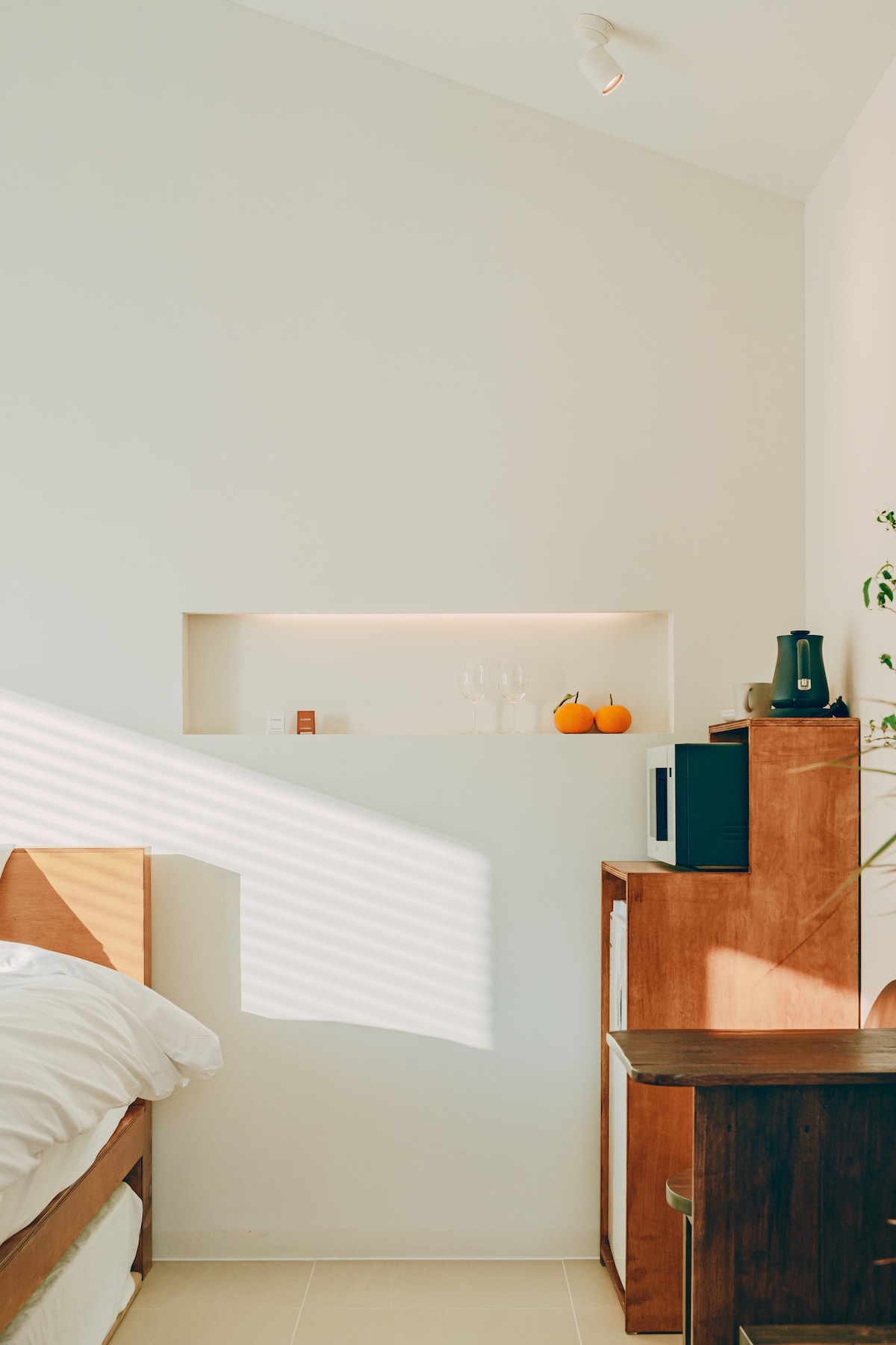 A bright and minimalistic interior is featured, showcasing a spacious bed and a wooden desk. A wall niche is illuminated, displaying two glasses and a small decorative pumpkin. A coffee maker and microwave are visible on the wooden cabinet, enhancing the room's functionality.