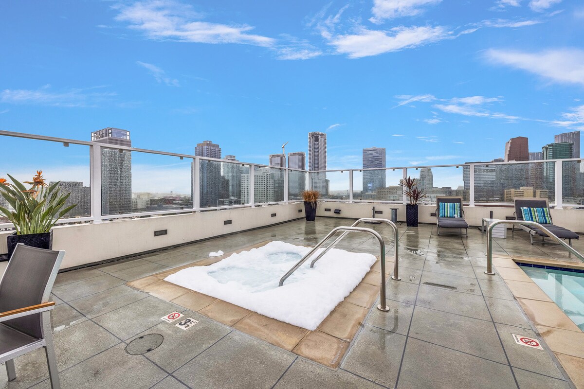 A hot tub is positioned on a tiled rooftop deck, surrounded by city views and greenery. Lounge chairs are available for relaxation, while the skyline of tall buildings stretches in the distance under a clear blue sky.