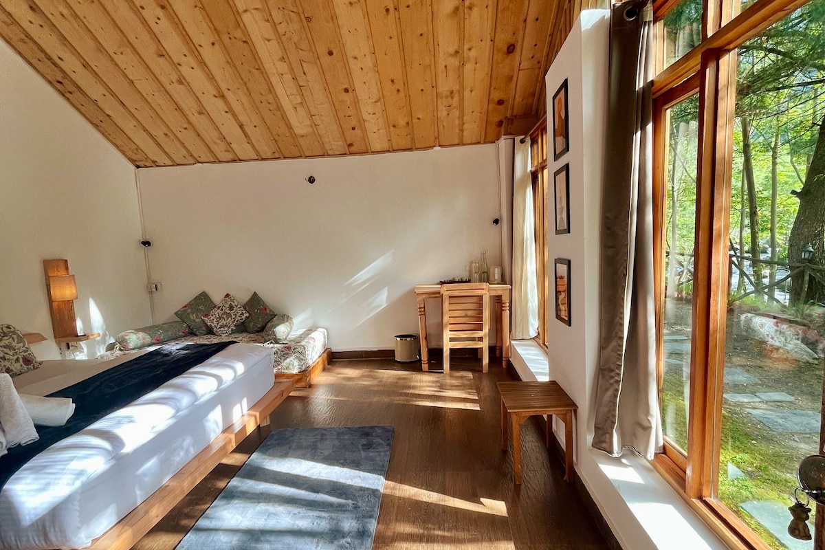 A serene room features a spacious bed, wooden ceiling, and large windows that allow natural light to fill the space. A small dining table with two chairs and a wooden bench are positioned near the windows, enhancing the room's inviting feel.