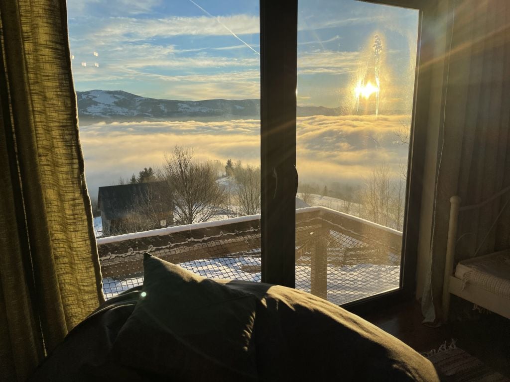 A large glass door offers a stunning view of a sun setting over a sea of clouds. The warm light fills the room, reflecting off the glass. Soft furnishings are positioned in front of the door, creating a cozy interior atmosphere.