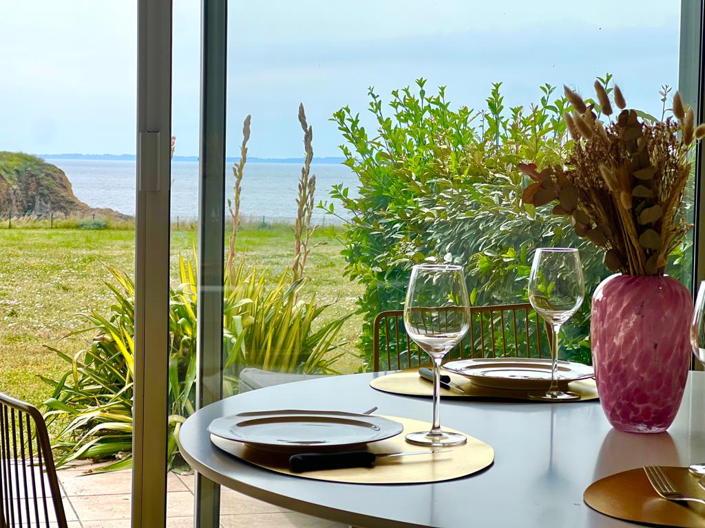 A dining area is presented with a round table set for two, embellished with elegant glassware and decorative dishes. A vibrant pink vase holds dried flowers, while a view of the ocean and lush greenery is framed through expansive glass doors.