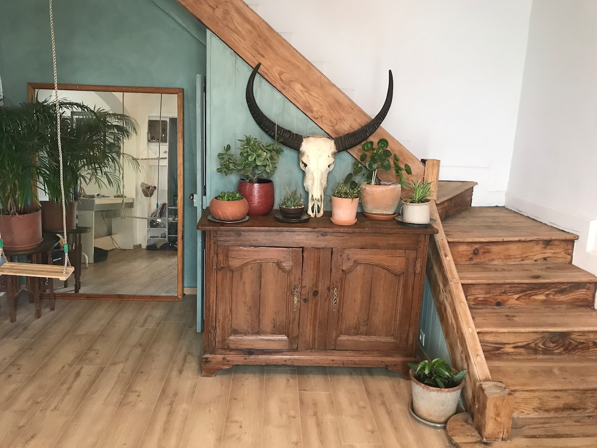 A wooden cabinet is positioned against a wall, adorned with various plants in pots. A decorative cow skull is mounted above the cabinet. The space features light-colored wooden flooring and a staircase made of natural wood, leading to the upper levels of the home.