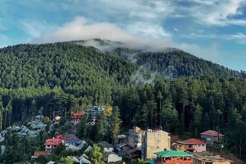 A scenic view captures lush green hills and cloud formations above, with a quaint village nestled among the trees. Colorful rooftops of nearby houses are visible, surrounded by dense forests, creating a serene landscape that invites relaxation.