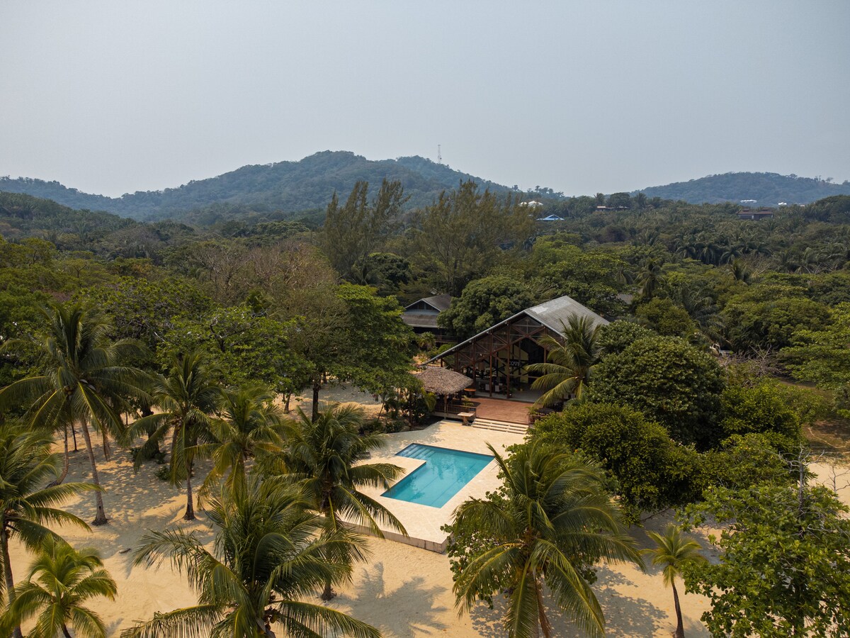 An aerial view of the villa reveals a spacious home surrounded by lush greenery and palm trees. A clean swimming pool is situated adjacent to the house, offering a bright blue contrast against the sandy landscape. The distant mountains provide a serene backdrop.