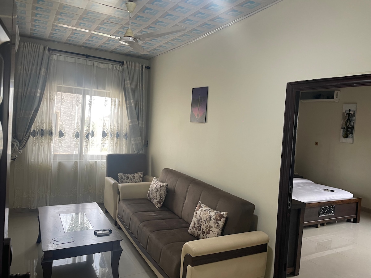 A comfortable living area is presented, featuring a well-upholstered sofa with decorative cushions. A wooden coffee table sits in front, with natural light brightening the space through large windows adorned with sheer curtains. A subtle piece of wall art adds a touch of elegance.