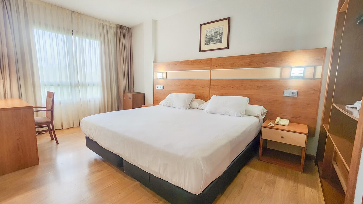 A spacious guest room is furnished with a double bed and a wooden desk with a chair. Natural light filters through large curtains, illuminating the light wood flooring. Wall-mounted lamps provide soft lighting, while a piece of artwork adorns the wall above the bed.