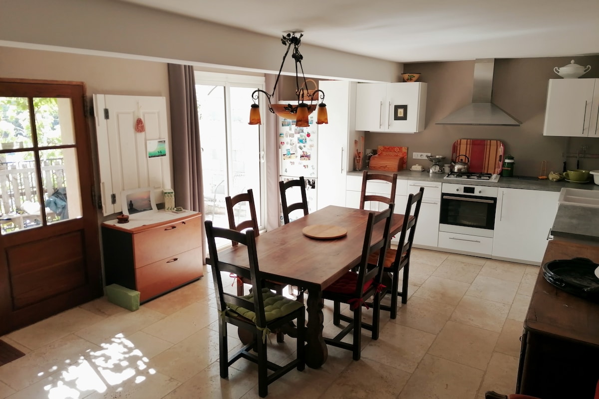 A dining area is featured with a wooden table surrounded by six chairs. A light fixture hangs overhead, while a kitchen with modern appliances is visible in the background. Natural light enters through the sliding doors, enhancing the warm tones of the space.