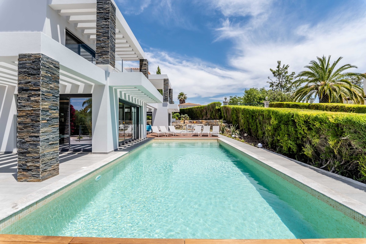 The image displays a well-maintained heated swimming pool, reflecting the clear blue sky. Surrounding the pool is a spacious deck furnished with sun loungers for relaxation. The modern villa architecture features large glass doors, blending indoor and outdoor spaces seamlessly, with greenery visible in the background.