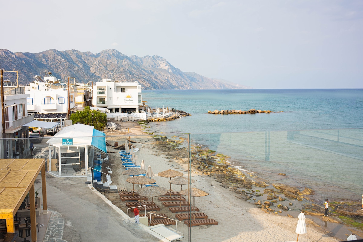 A view of a serene beach extends along the coastline, featuring clear waters and rocky outcrops. Sun loungers and umbrellas are arranged on the sandy shore, with a backdrop of low-rise buildings and distant mountains under a partially cloudy sky.