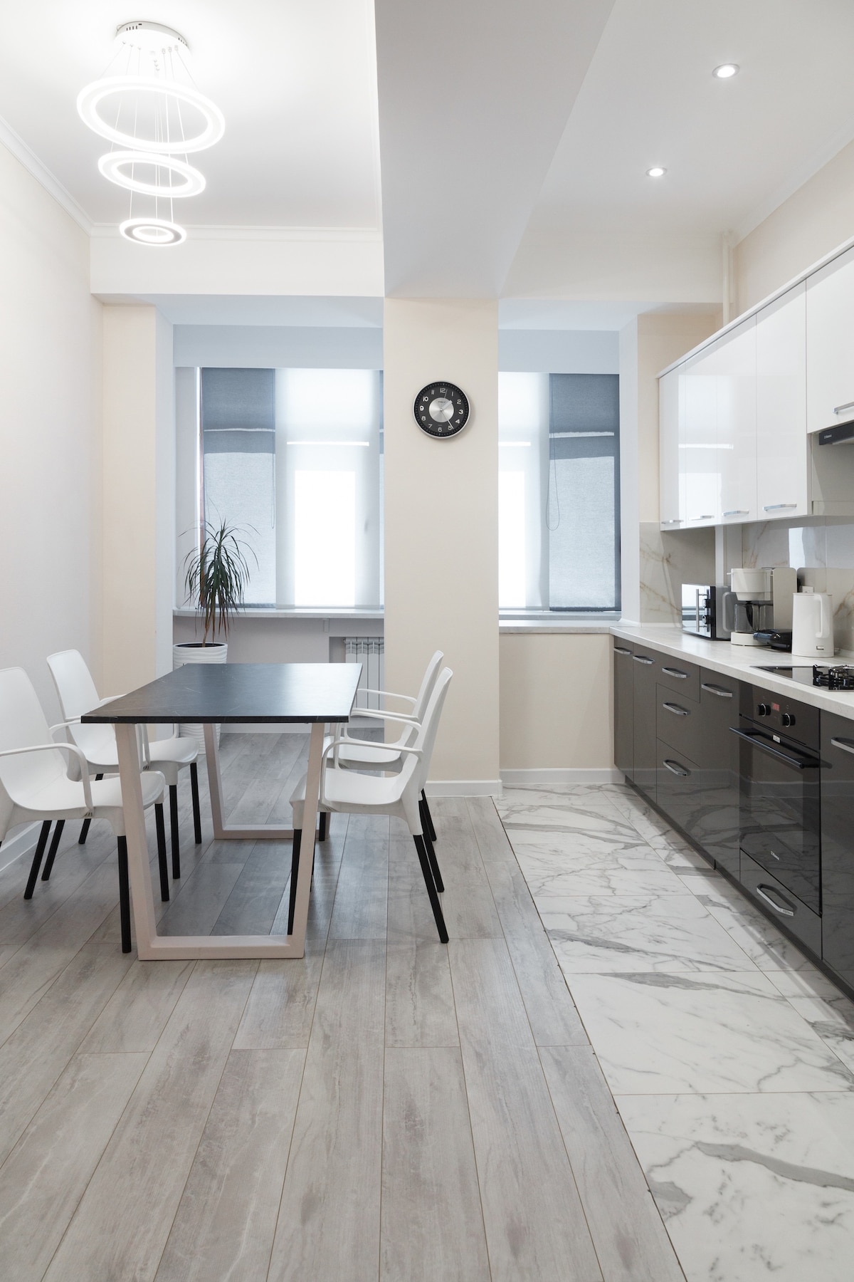 A modern kitchen area features sleek cabinetry and appliances, complemented by a minimalist dining table surrounded by white chairs. Natural light enters through large windows, creating a bright ambience. The floor alternates between light gray wood and elegant marble tiles for added visual interest.