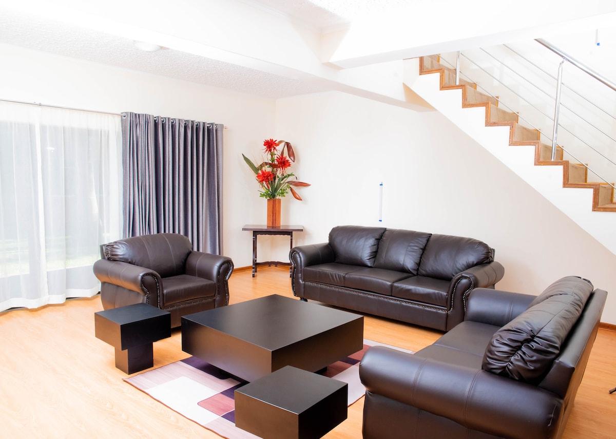 A spacious living area features dark leather sofas arranged around a black coffee table. A striped rug adds a touch of comfort to the wood floor. Natural light filters through sheer curtains, while vibrant plants are placed on a nearby table, enhancing the inviting atmosphere.