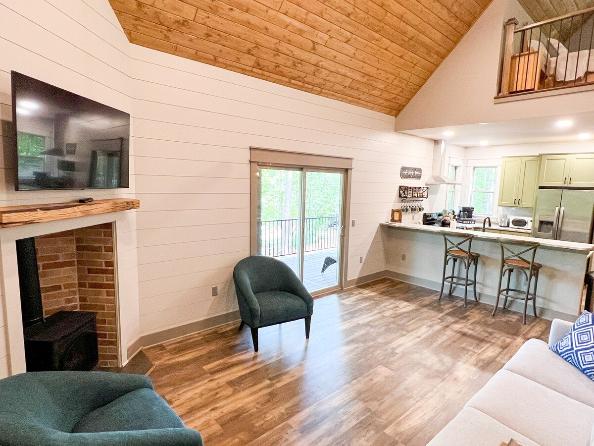 An open living area is featured, showcasing a comfortable seating arrangement with two green chairs and a beige sofa. A television is mounted on the wall above a rustic fireplace. Light enters through sliding doors that lead to a balcony, enhancing the airy space.