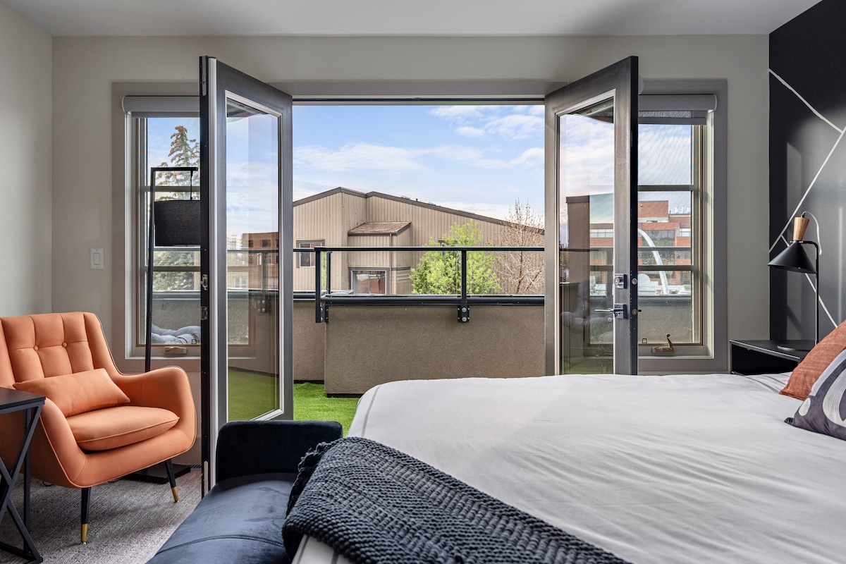 The master bedroom features an open balcony with a view of nearby buildings and greenery. Large glass doors allow natural light to fill the space. A spacious bed is complemented by a comfortable orange armchair and a dark side table.