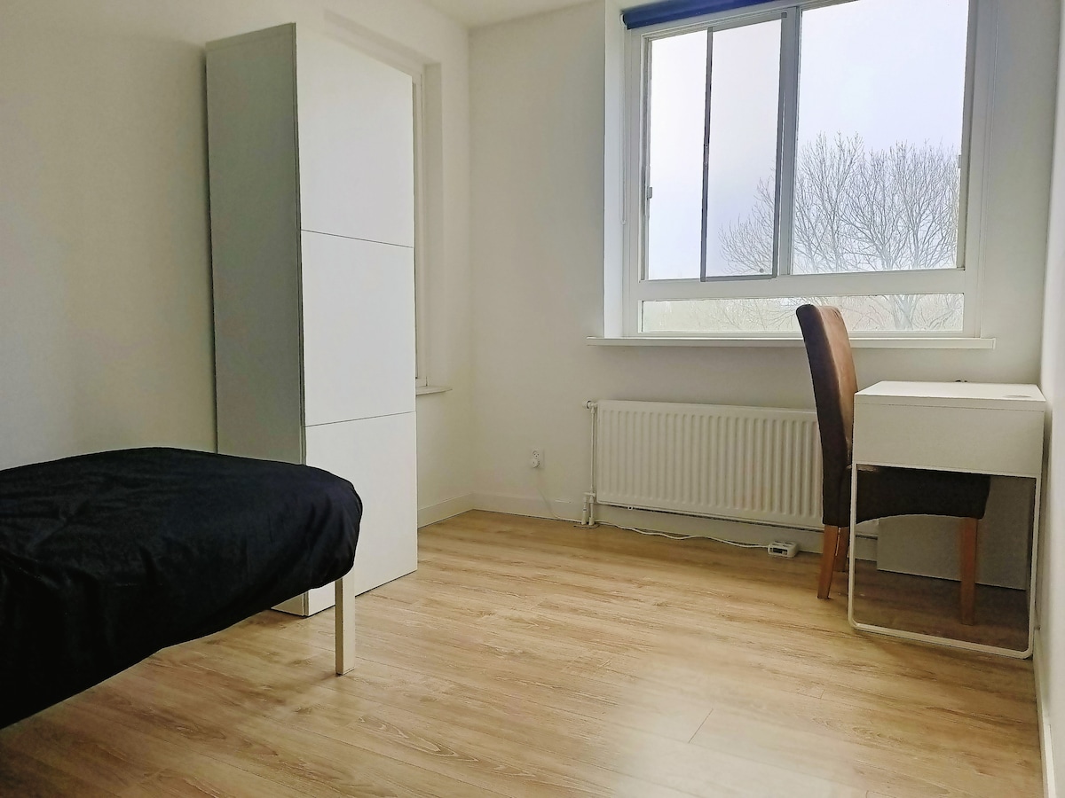 A bright room features a spacious bed with a dark cover, a modern wardrobe against the wall, and a desk with a chair. Natural light comes through a large window, providing a view of bare trees outside.