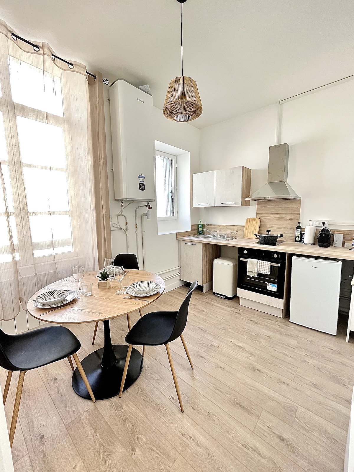A modern kitchen is shown, featuring a round dining table with two black chairs, elegantly set with dishes. Light wood cabinets and countertops are complemented by a compact white refrigerator and an electric stove. Natural light floods in through a nearby window, highlighting the clean design.