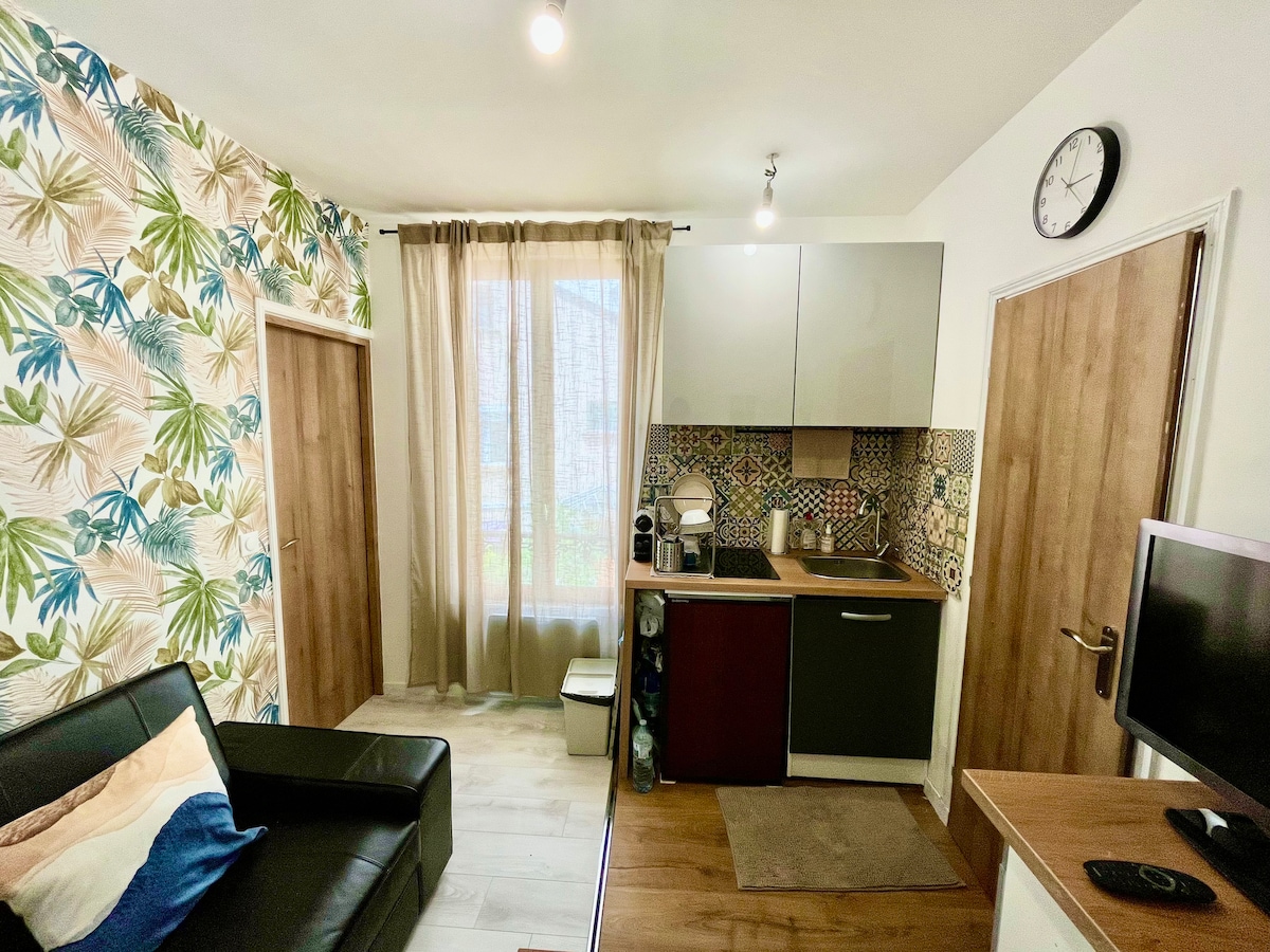 A modern living space features a black leather sofa, a compact kitchen with a patterned backsplash and essential appliances. Natural light floods through the window, which is adorned with sheer curtains. A wall clock is mounted for convenience, enhancing the functionality of the area.