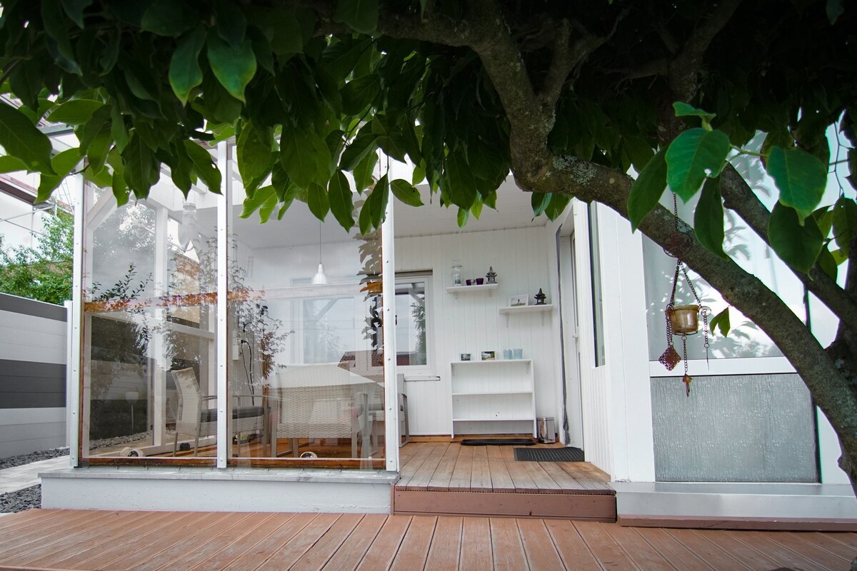A spacious outdoor area is framed by lush greenery, featuring large glass doors that open into a bright interior. The wooden deck extends towards a cozy seating area, and the gentle dappled light creates a serene atmosphere.