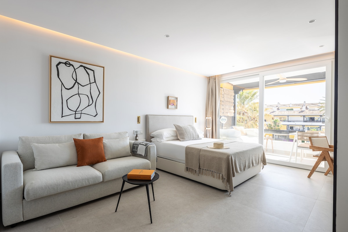 A modern studio space features a queen bed and a convertible sofa. Natural light flows in through large glass doors that open to a balcony. Minimalist artwork adorns the wall, and a small circular table sits beside the sofa, creating a comfortable living area.