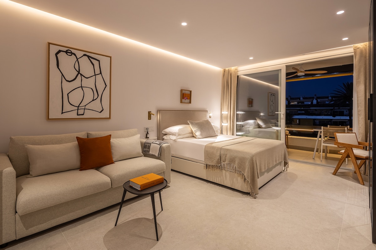 A modern studio layout features a comfortable Queen bed and a sofa bed, with a light-toned color palette creating a calm atmosphere. A balcony door offers views, and a small table and chair provide a workspace. Soft lighting highlights the minimalist decor.