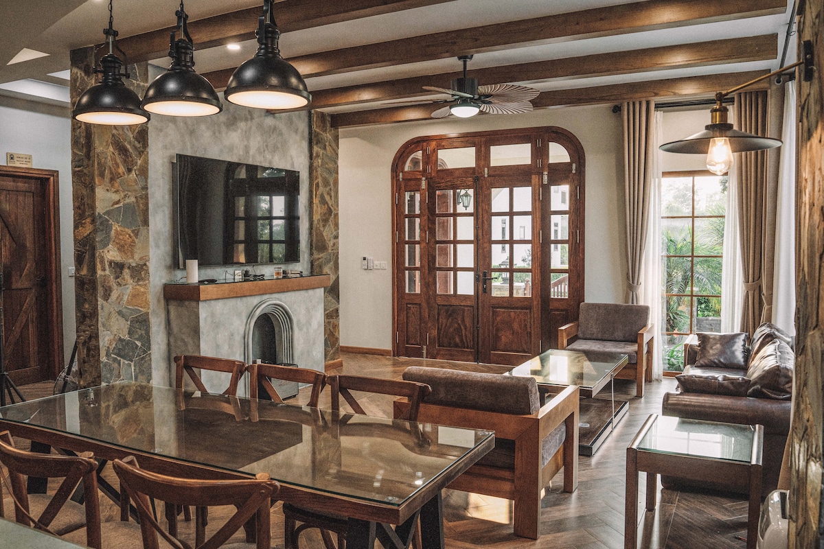 A spacious living and dining area is depicted, featuring a wooden dining table surrounded by chairs. A large flat-screen TV is mounted on the stone wall, complemented by natural light filtering through large windows with wooden frames.
