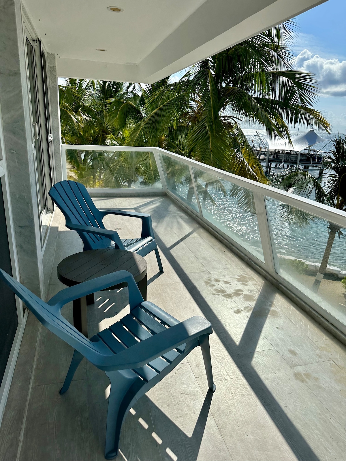 A private balcony presents two blue chairs and a small black table, overlooking the tranquil water and palm trees. The view captures the coastal scenery and showcases the bright sky, inviting relaxation in a serene atmosphere.