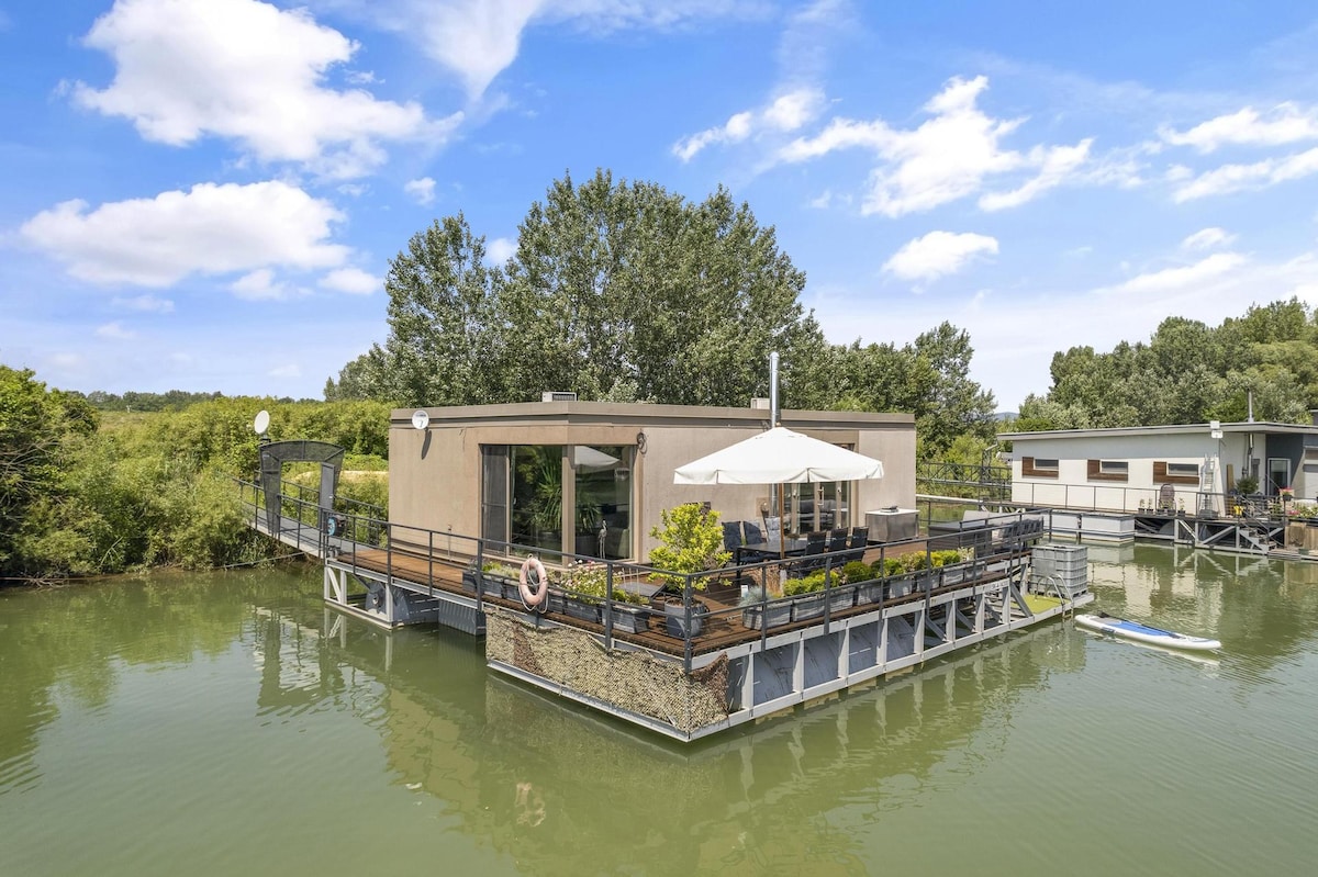A modern floating home is situated on calm waters, surrounded by lush greenery. A spacious outdoor terrace is equipped with a sunshade and adorned with plants. Surrounding pathways offer access to the structure, while a neighboring houseboat appears in the background.