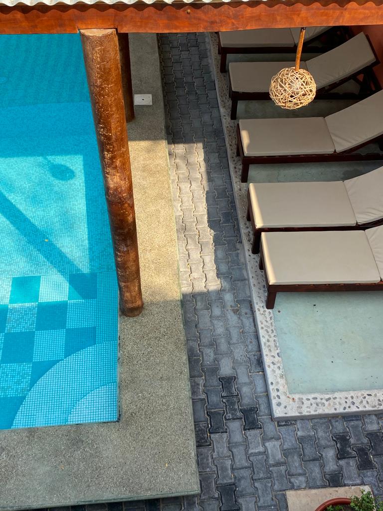 The image captures a tranquil pool area featuring a clear blue water surface adjacent to a shaded seating area with four lounge chairs. The flooring consists of a textured stone pathway, while a decorative light fixture is hung above, enhancing the serene outdoor setting.