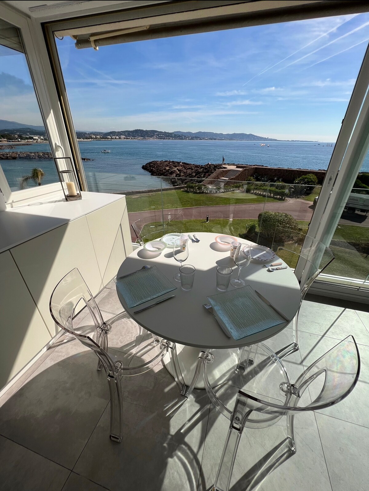 A modern dining area features a round table set for four, with clear acrylic chairs positioned around it. A stunning view of the bay and coastline can be seen through large windows, allowing natural light to brighten the space.