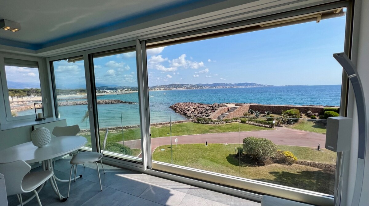 A panoramic view of the azure sea is framed by large glass windows, offering a serene backdrop. A round dining table with white chairs is positioned nearby, creating a comfortable space to enjoy the scenery. The coastline and distant hills are visible, contributing to the coastal atmosphere.