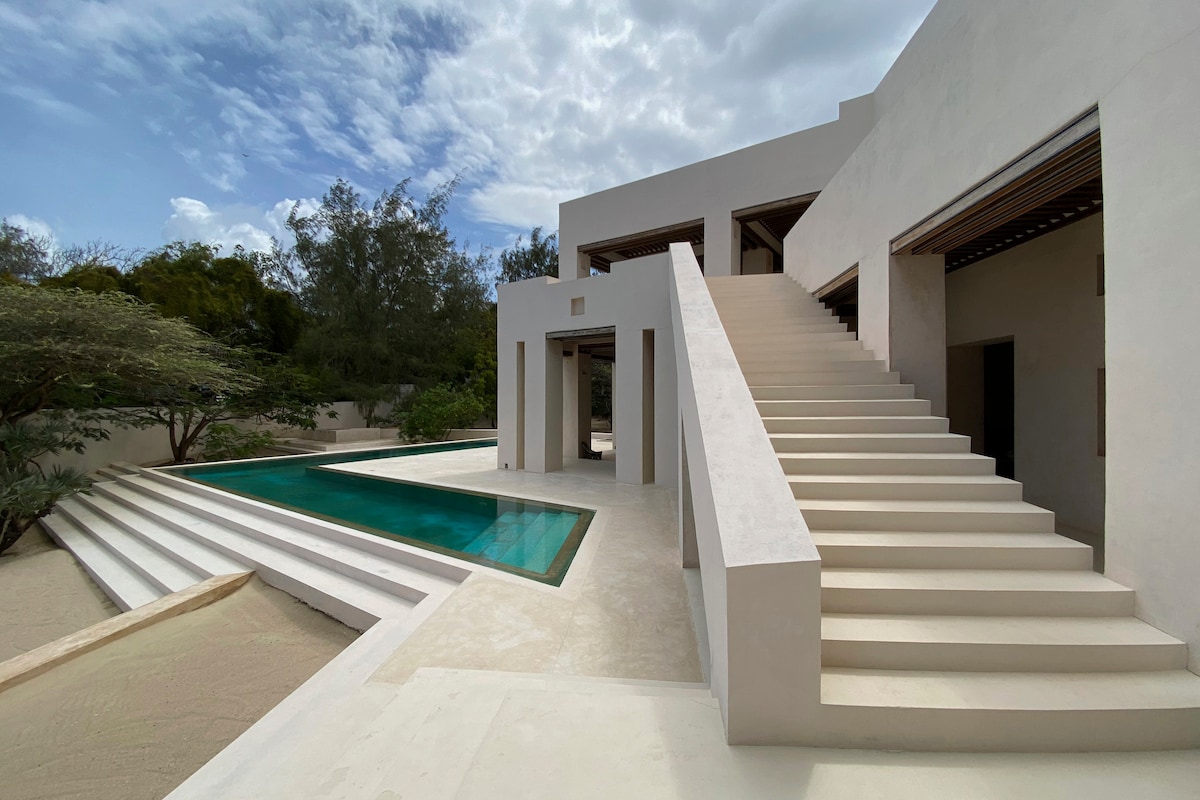 A modern home features a sleek, minimalist staircase leading up to an upper level. The spacious pool reflects the sky, with surrounding steps providing easy access. Lush greenery is visible around the property, enhancing the serene outdoor environment.