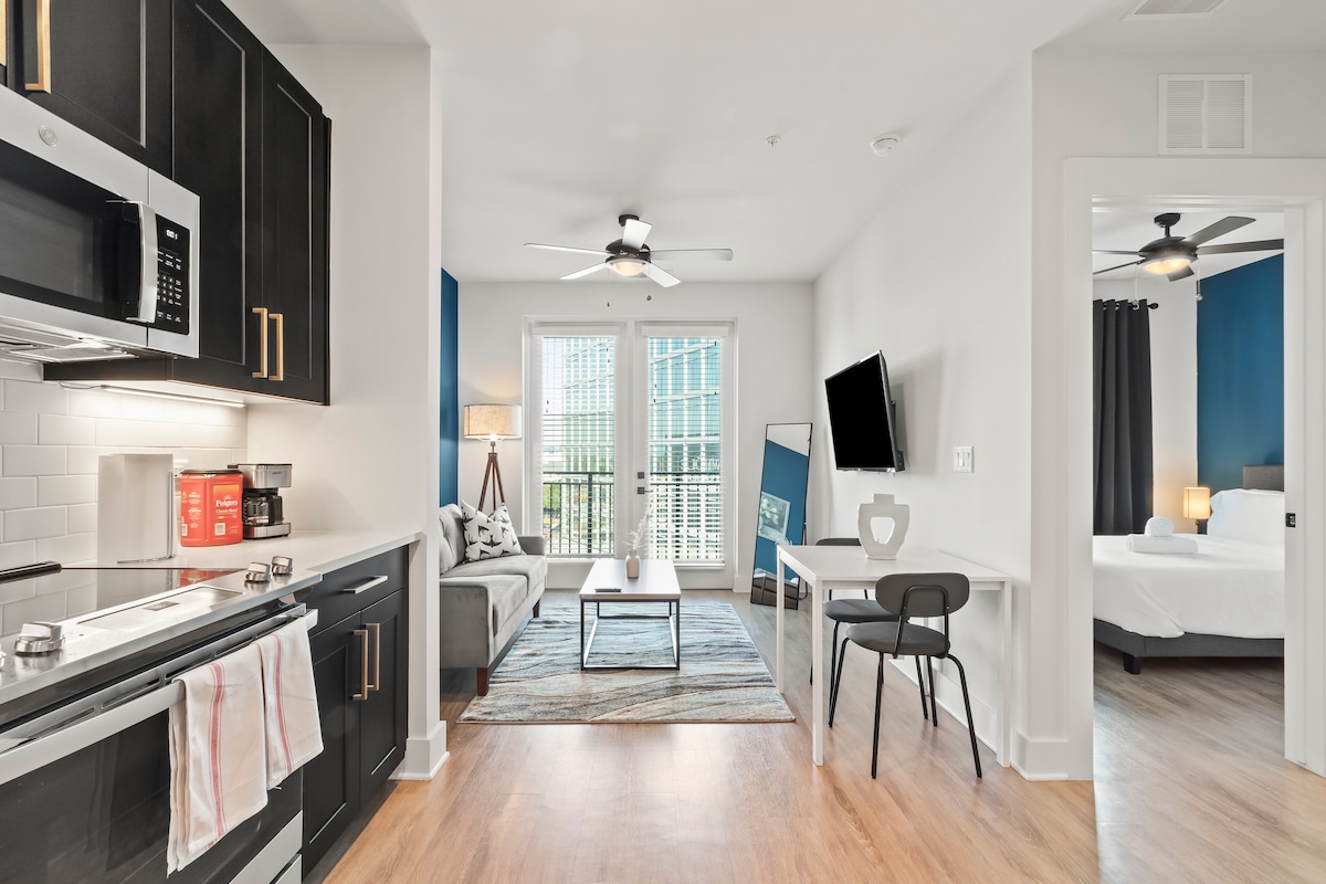 The studio features a modern kitchen with stainless steel appliances and a cozy living area furnished with a sofa and a coffee table. Natural light fills the space from large windows, enhancing a relaxed ambiance. A dining table and wall-mounted TV are also visible.