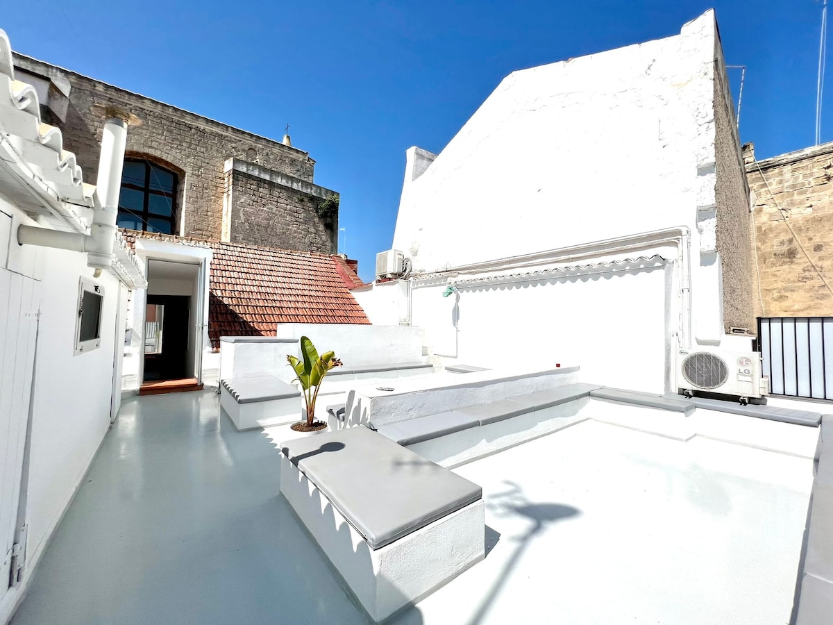 A spacious rooftop terrace is presented with a clean, white finish. Potted greenery adds a touch of nature. An area is designated for lounging, and cooling units are visible along the walls. The bright blue sky provides ample natural light to the space.