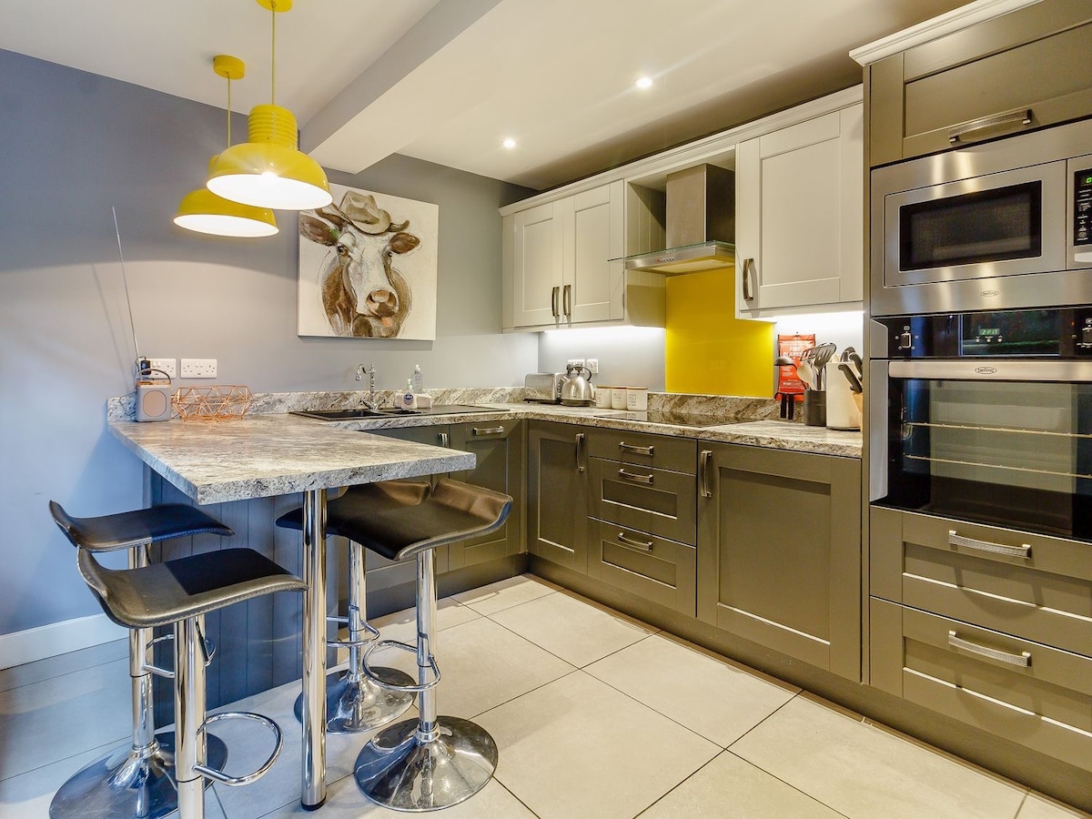A compact kitchen is equipped with gray cabinetry and modern appliances, including an oven and microwave. A breakfast bar with four high stools offers a casual dining option. Bright yellow pendant lights hang above, illuminating the stone countertop and the artwork of a cow on the wall.