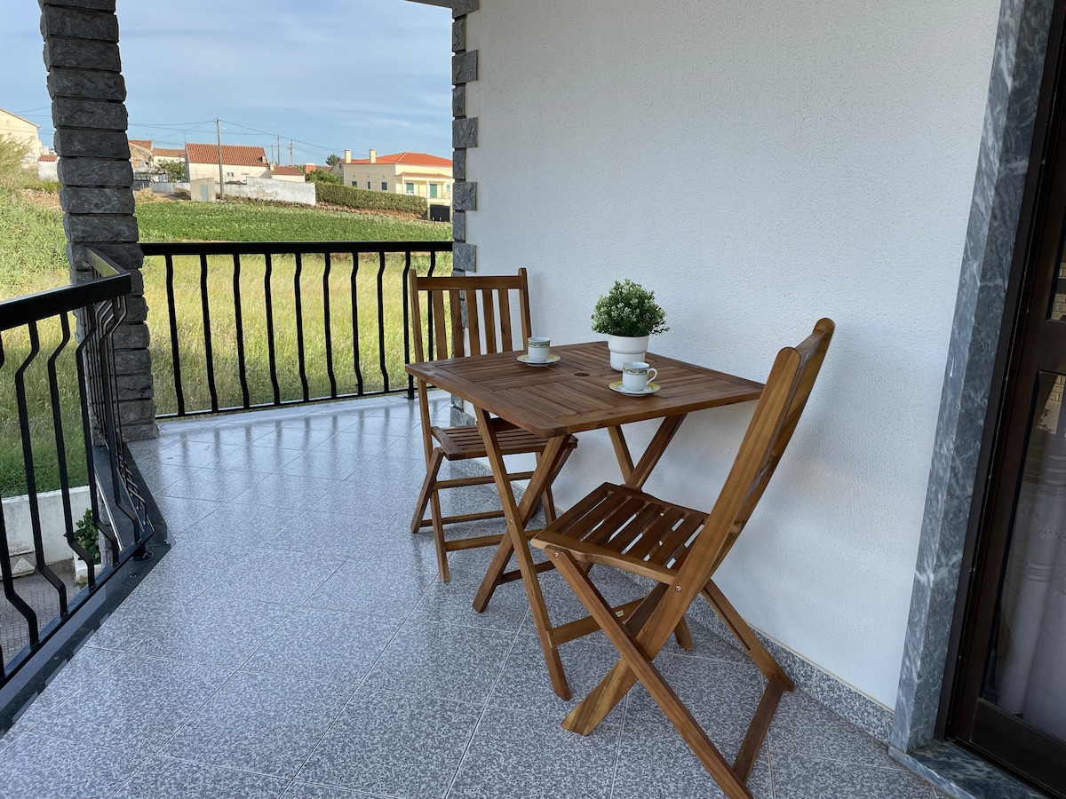 A wooden table is set with two cups, accompanied by two folding chairs on a covered terrace. The area features a smooth tiled floor and overlooks a grassy field, providing a serene view of the surrounding landscape.