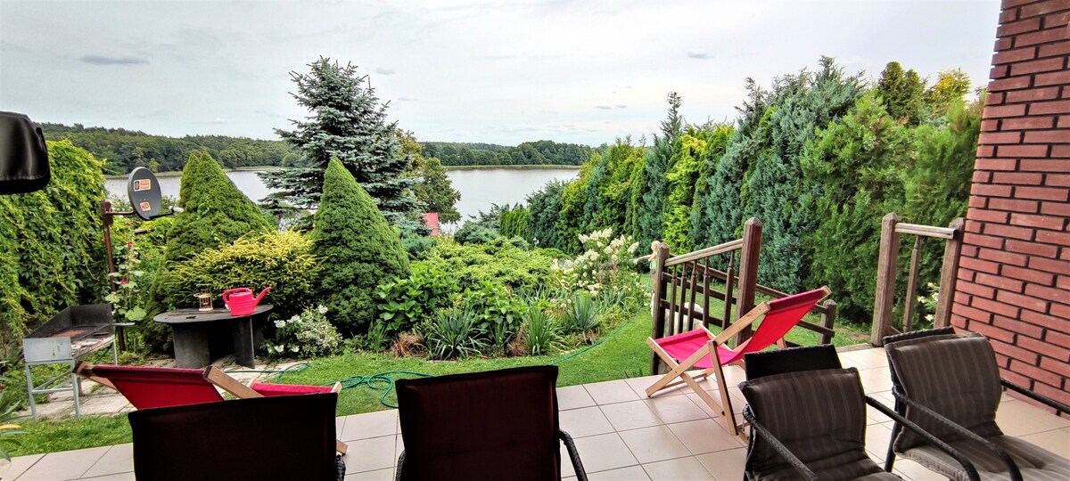 An outdoor patio features comfortable seating arrangements, including lounge chairs and recliners. Lush greenery surrounds the area, leading to a serene lake view in the background. A grill is positioned nearby, and pathways through the garden enhance the natural setting.