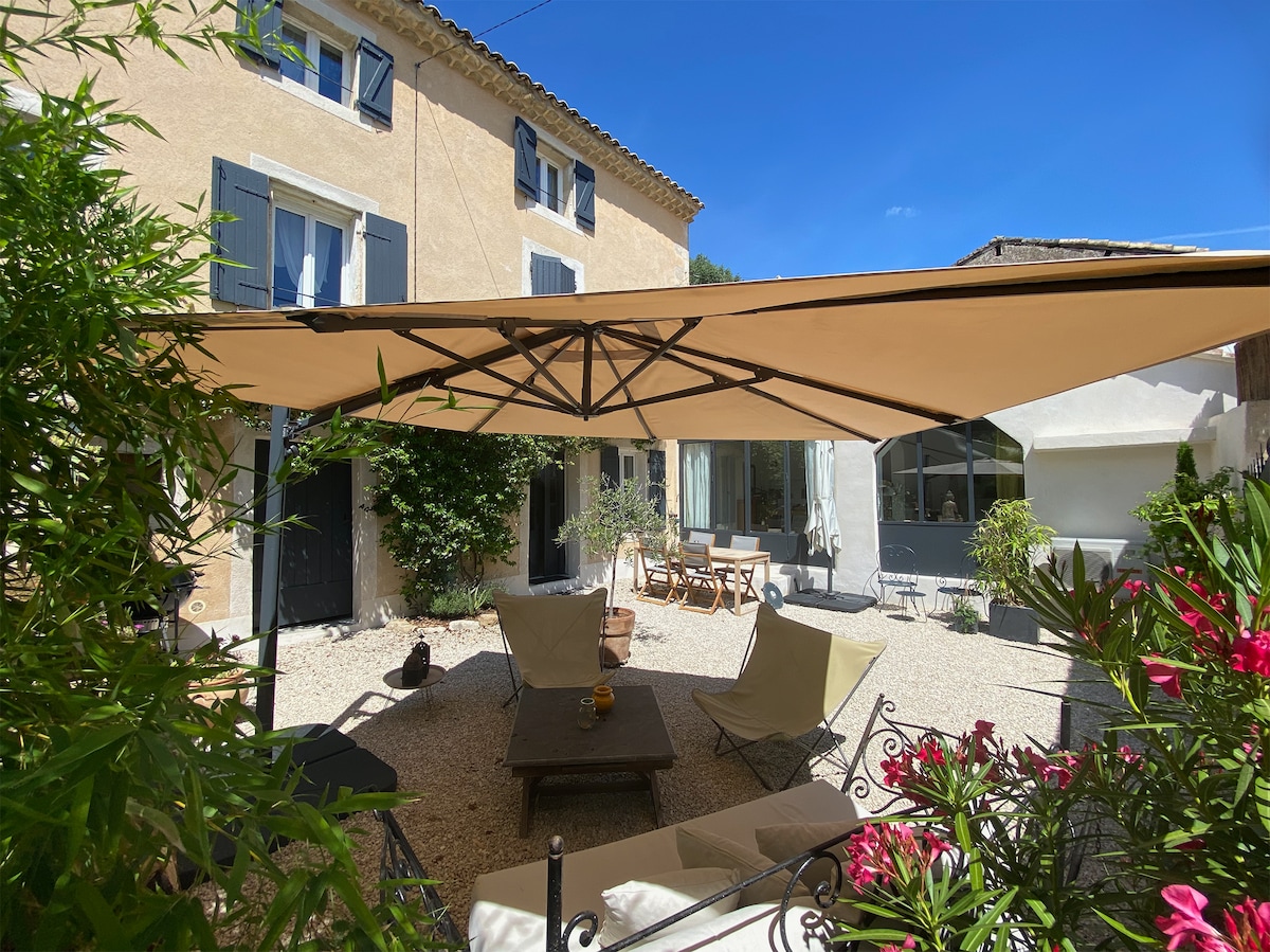 A shaded outdoor terrace is featured, adorned with comfortable seating and a coffee table. Lush greenery and colorful flowers enhance the space, creating a peaceful environment. The house's exterior, characterized by rustic architecture and blue shutters, is visible in the background, completing the inviting atmosphere.