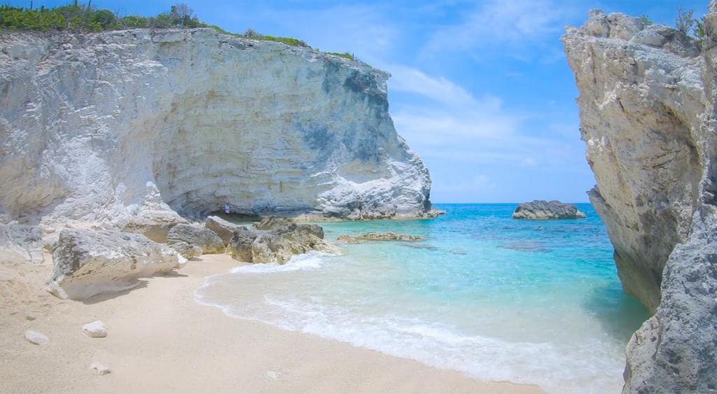 A serene beach scene features soft, white sand meeting crystal-clear turquoise waters. The coastline is framed by rugged, pale cliffs on either side, creating a natural and tranquil environment. A clear blue sky adds a sense of openness and calm to the landscape.