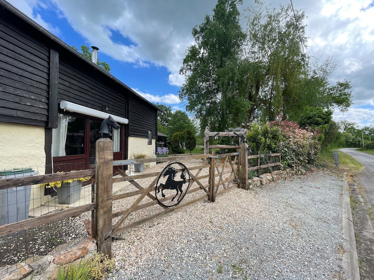 The exterior of a rustic property is framed by a wooden fence featuring a horse emblem. A gravel pathway leads to the entrance, flanked by greenery and flowering shrubs, with an open sky visible above.