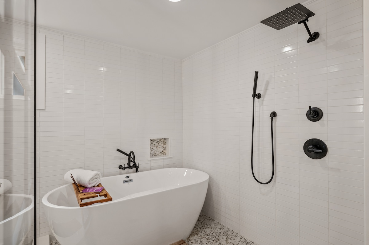 A modern bathroom features a freestanding soaking tub and a walk-in shower highlighted by a rainfall showerhead. White tile surrounds the relaxing space, with a towel neatly placed on the tub's edge and minimalist black fixtures enhancing the serene atmosphere.