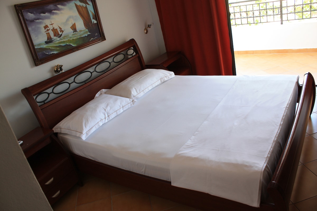 A spacious bed with crisp white linens is positioned beside a wooden nightstand. A nautical-themed painting adorns the wall, while light from a partially open window casts a soft glow on the tiled floor. Dark curtains provide an option for privacy.