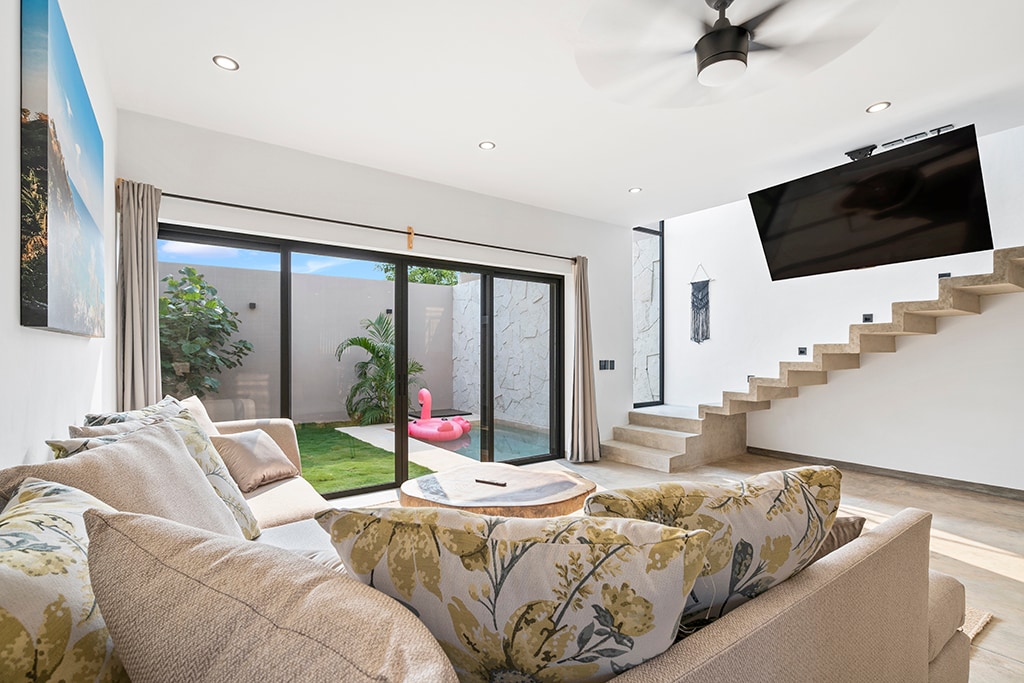 A cozy living area features a plush sectional sofa adorned with floral cushions. Large glass sliding doors open to a private courtyard with greenery. A wall-mounted TV is positioned above a staircase leading to the upper level, while a ceiling fan provides airflow.