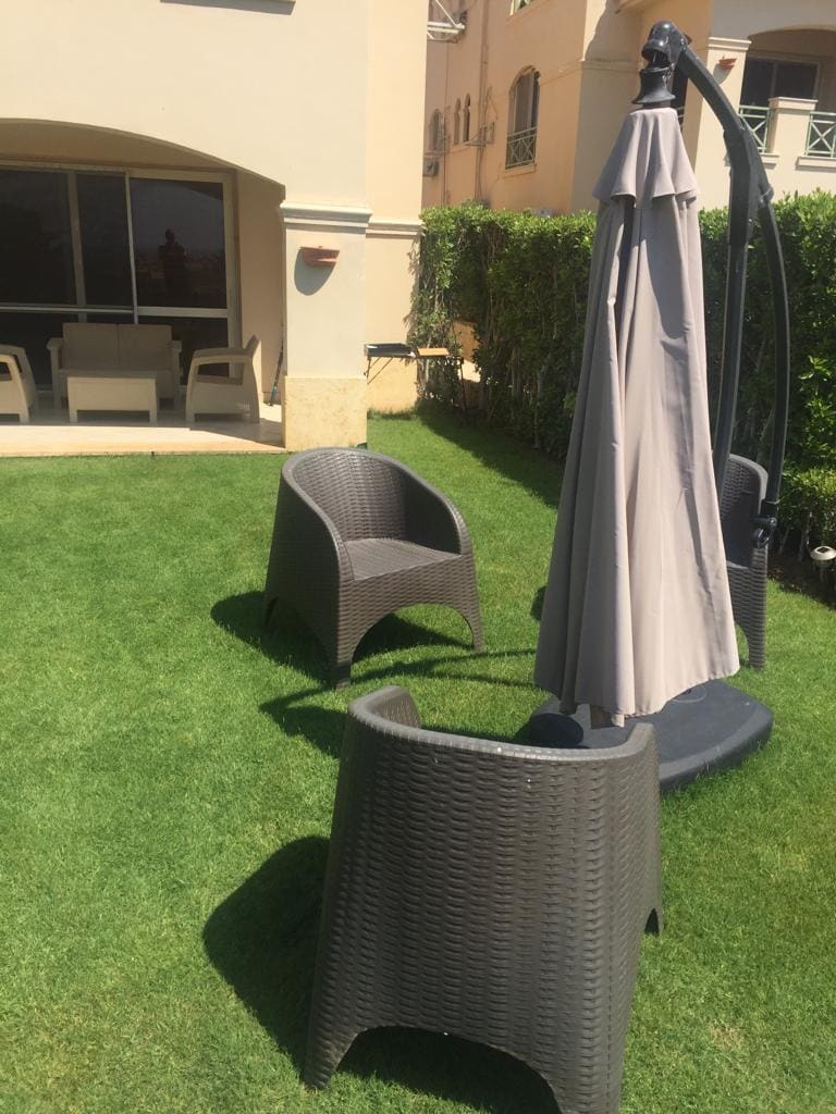 An outdoor space features well-maintained green grass, complemented by two stylish wicker chairs. A beige umbrella is positioned over a sturdy base, offering shade. In the background, a covered seating area can be seen through large glass doors.