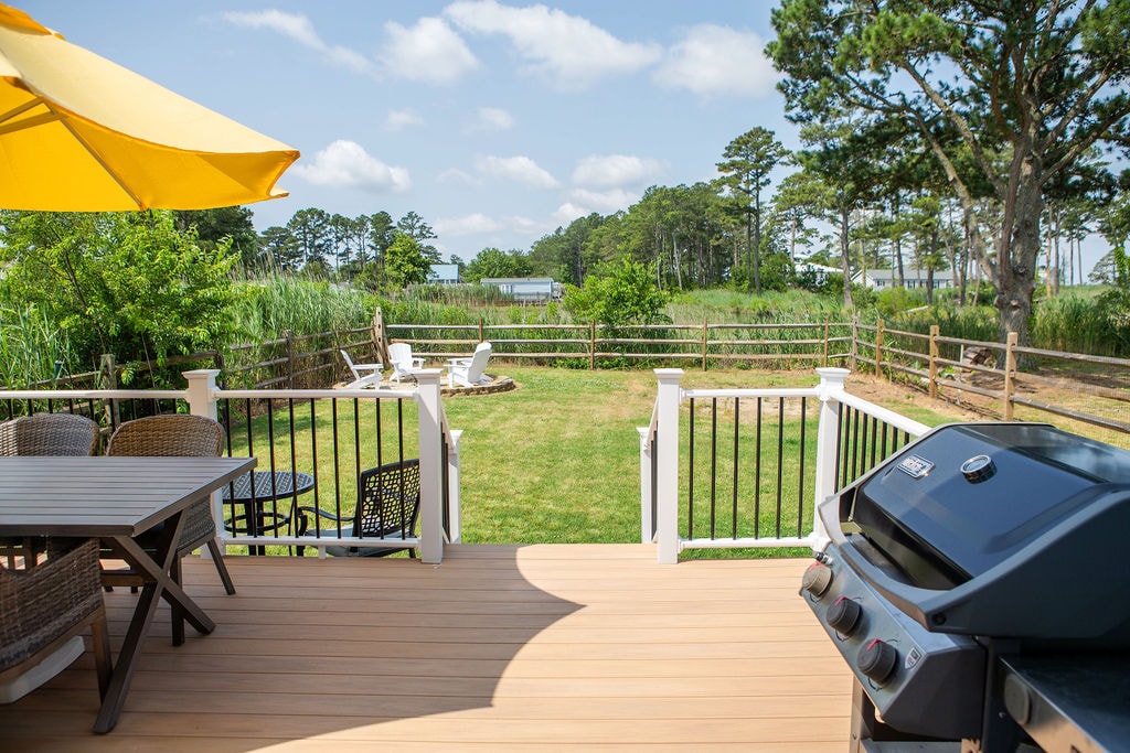 A spacious deck features a propane grill and a dining table with six chairs under a yellow umbrella. The fenced yard is visible beyond the deck, adorned with lush greenery and a fire pit surrounded by Adirondack chairs, offering a peaceful outdoor setting.