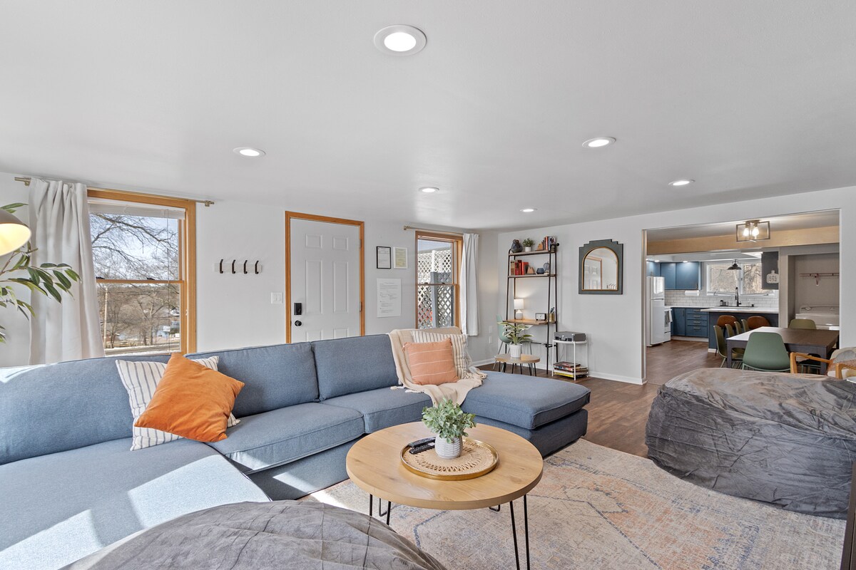 A spacious living room features a large sectional sofa adorned with colorful pillows. A circular coffee table holds a small plant, and light streams through multiple windows. The open layout connects to a dining area and kitchen, creating an inviting atmosphere for relaxation and gatherings.