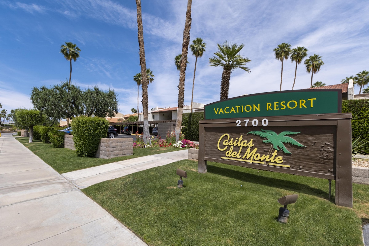 A welcoming entrance sign displays the name of Casitas del Monte, surrounded by manicured grass and palm trees. The pathway is lined with vibrant flowers, leading to the resort's main building visible in the background, enhancing the serene desert atmosphere.